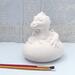 Ceramic baby dragone to paint sitting on a blue table with a lacy ecru curtain behind it.  There are two used paint brushes in front of it.